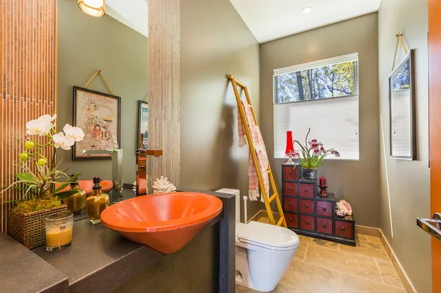 An orange basin sits among warm textured surfaces, plants and a leaning mirror.
