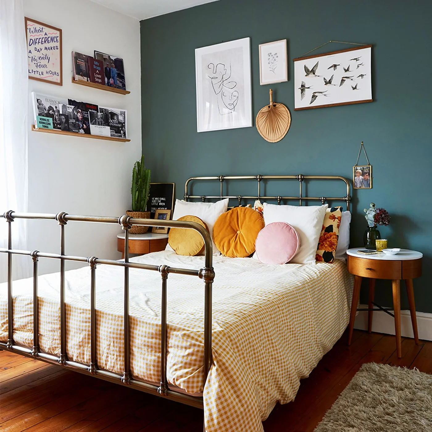 A dark green wall with mixed framed pictures sits behind an open metal bed with white bedding, warm-colored cushions and round bedside tables