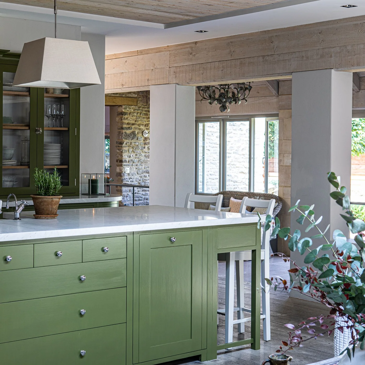 Olive green island drawers and glass-fronted storage sit beneath broad wooden beams beside a pale counter and white stools