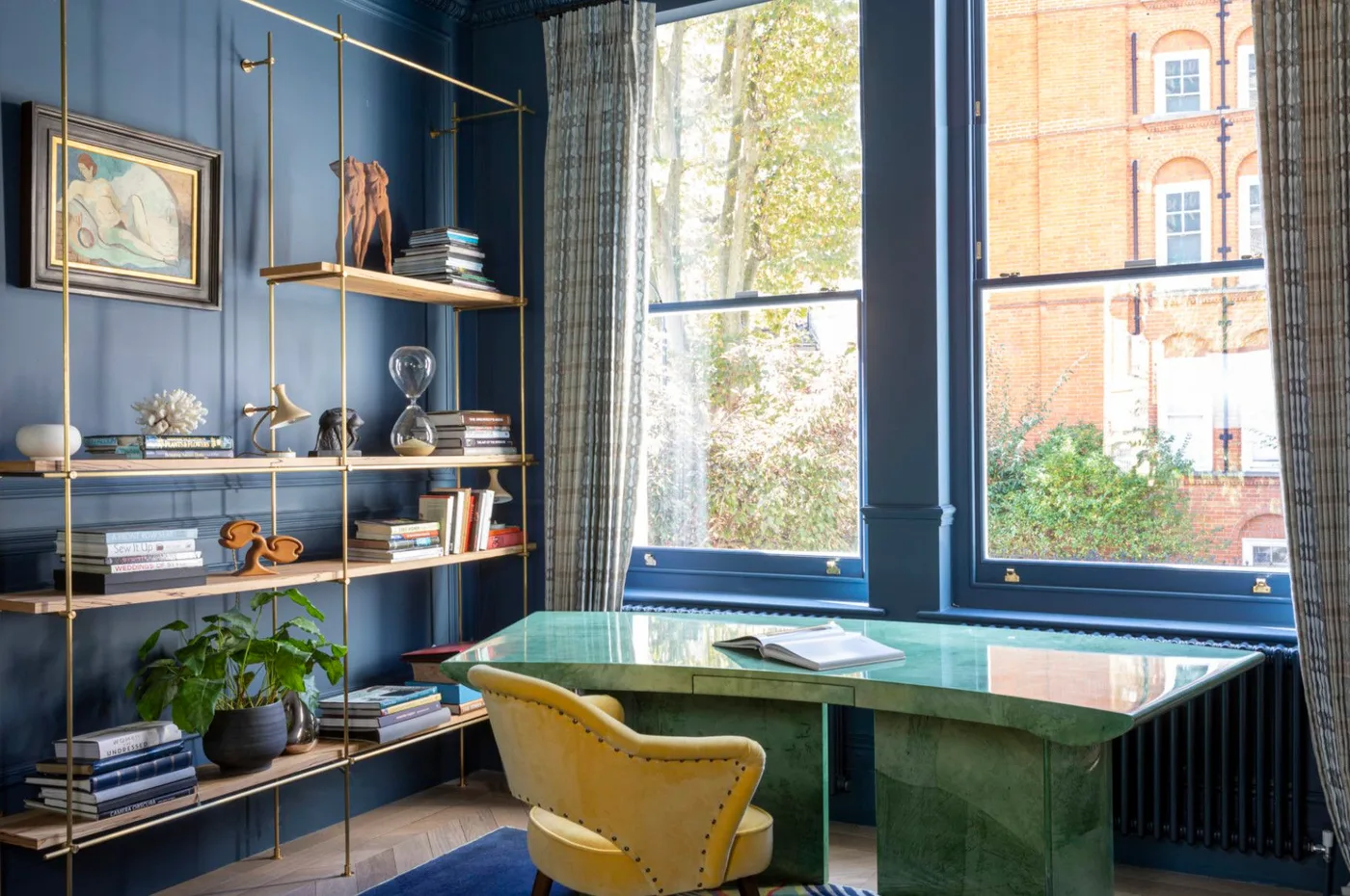 A glossy green desk with a yellow chair sits before blue walls beside warm metal open shelving and tall windows