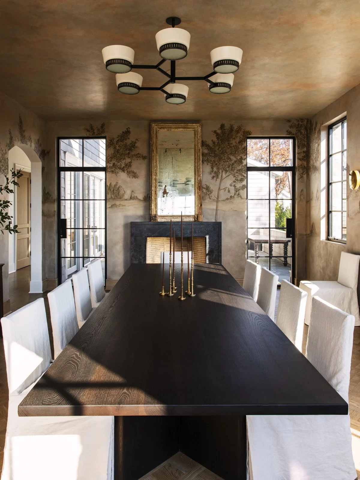 A long dark dining table sits between pale upholstered chairs beneath a multi-shade ceiling light with scenic walls, a fireplace and tall framed mirror