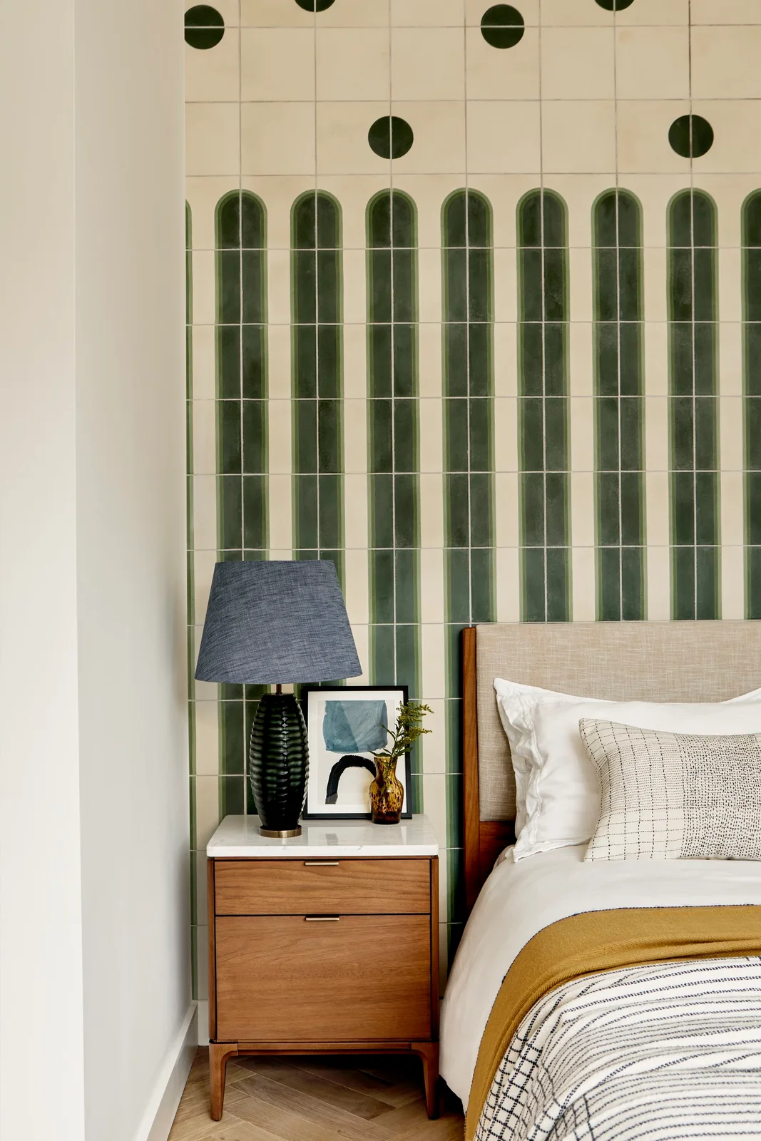 Tall green tile-like shapes and dark circles repeat on a pale bedroom wall behind a simple upholstered headboard beside a wood cabinet and dark lamp