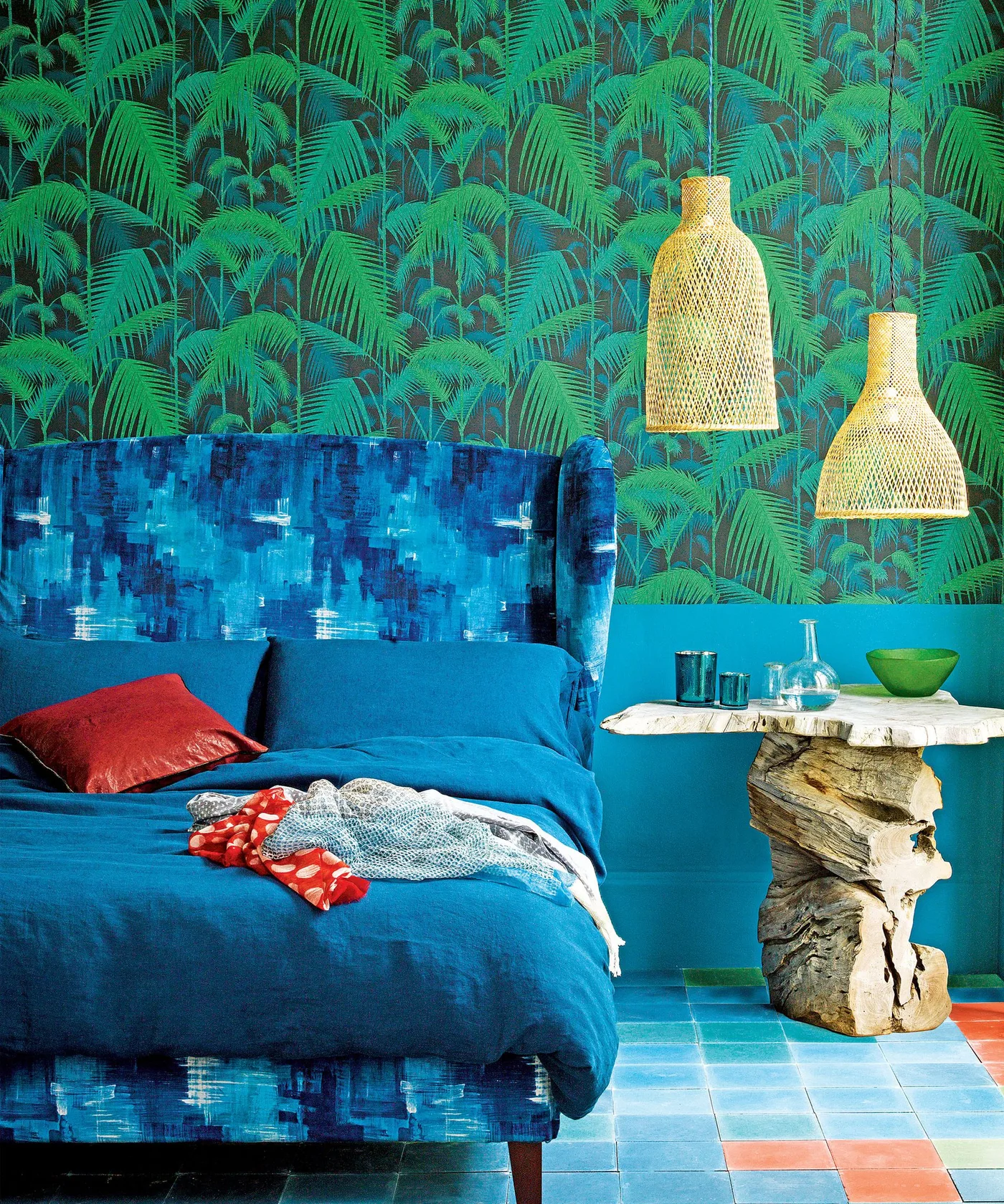 Bright green palm wallpaper above a blue upholstered bed with woven pendant shades and a sculptural side table.