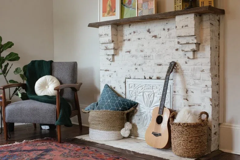 Partly white-coated brick fireplace with a dark wooden mantel, baskets and a guitar in front of the opening.