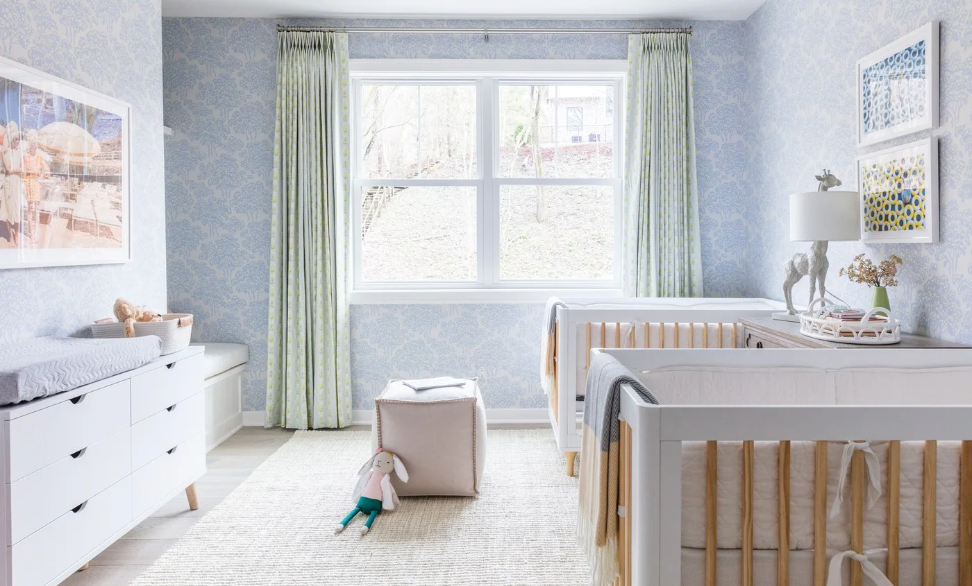 Two pale nursery cribs with wood slats stand beside white drawer storage, pale blue patterned walls, green curtains and a small rounded footrest