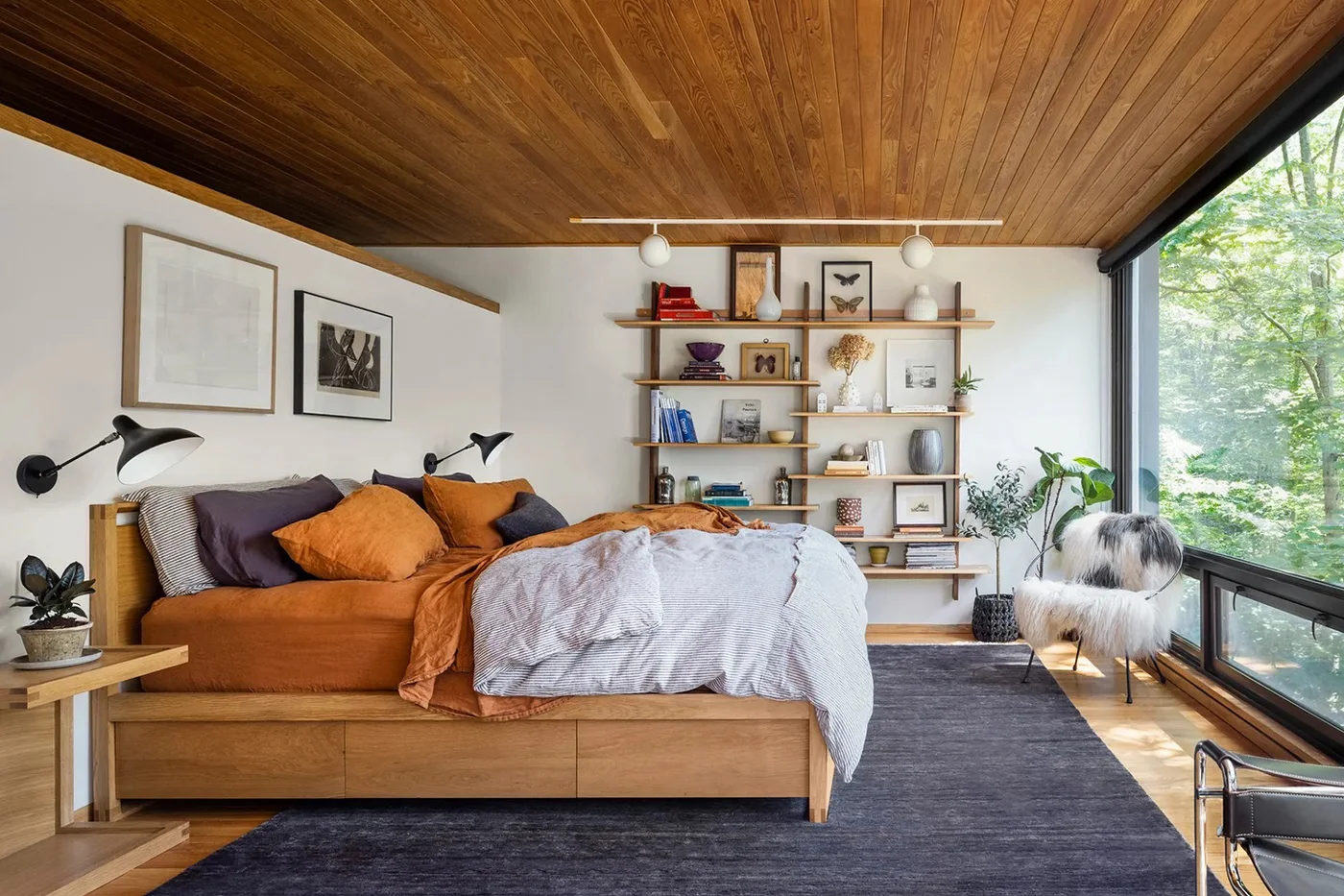 A wood platform bed with orange and gray bedding sits beneath a wood ceiling beside open wall shelves, black reading lights and large garden-facing glazing