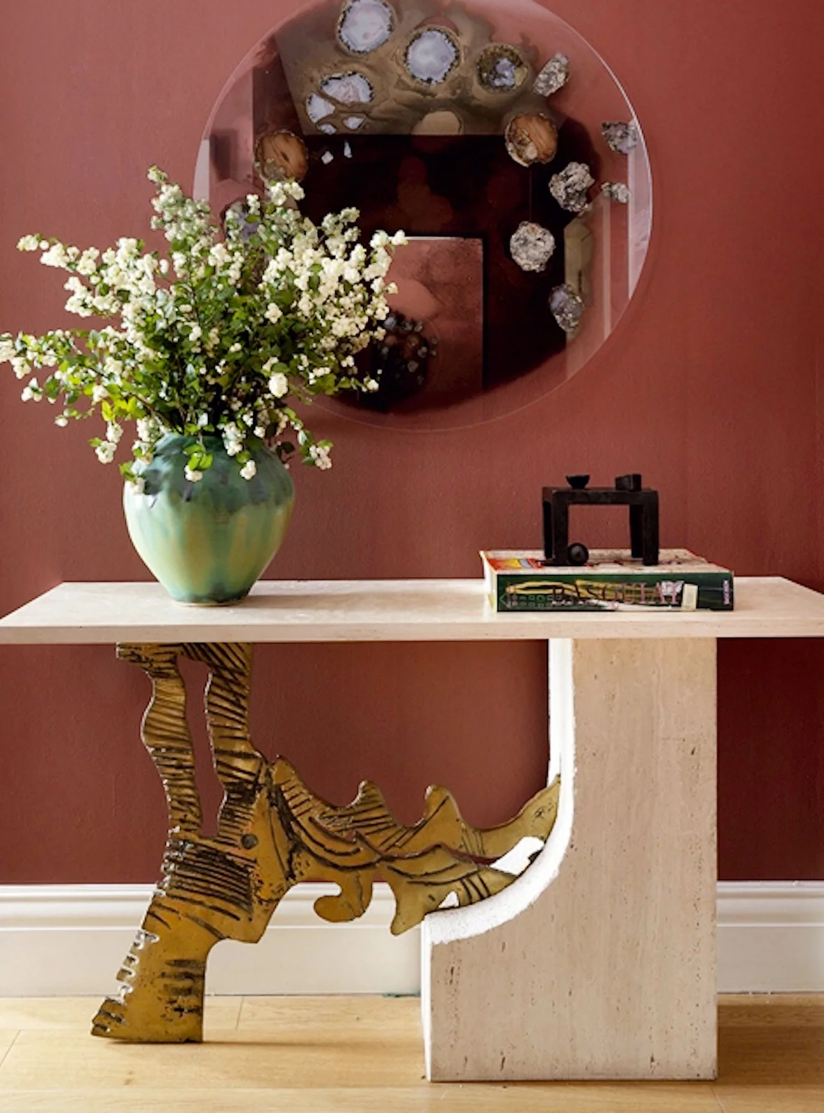 A pale curved console stands against a pink wall beneath a round mirror with flowers above and a striped tiger sculpture underneath