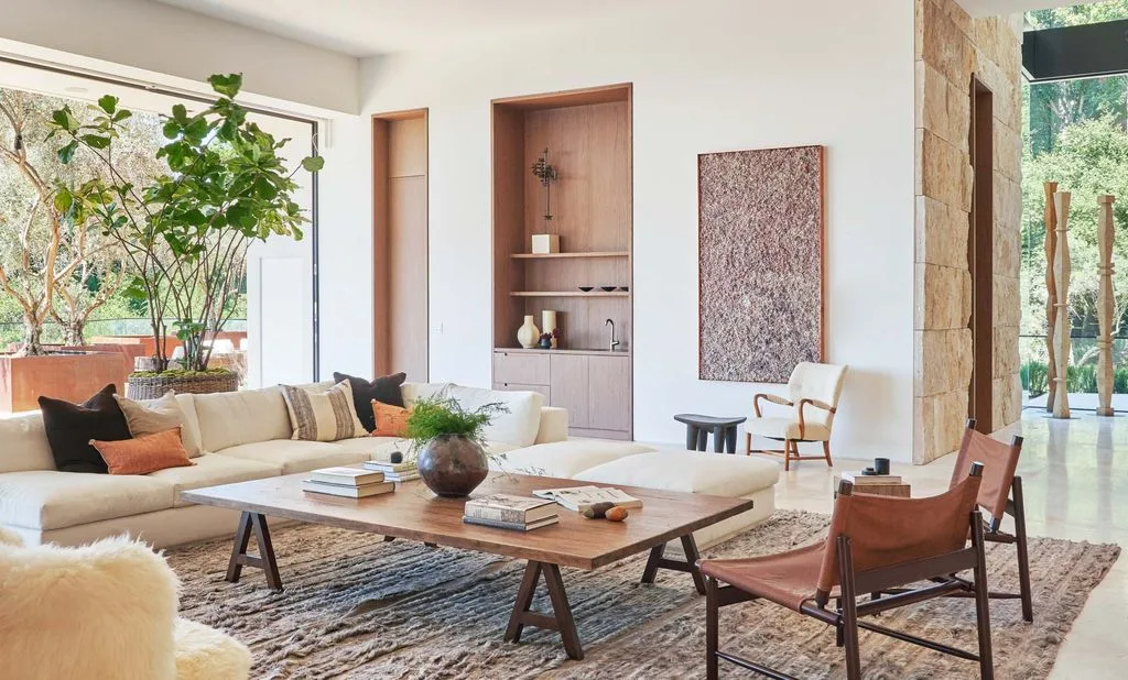 Pale living room with a low sectional sofa, wooden coffee table, warm-colored chairs, recessed shelves and a textured rug.