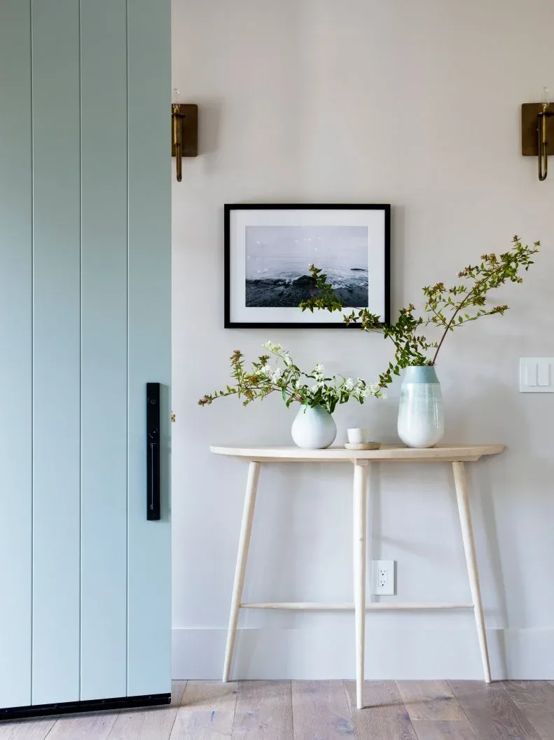 Pale wood console beneath framed landscape beside pale blue boarded front door and wall sconces