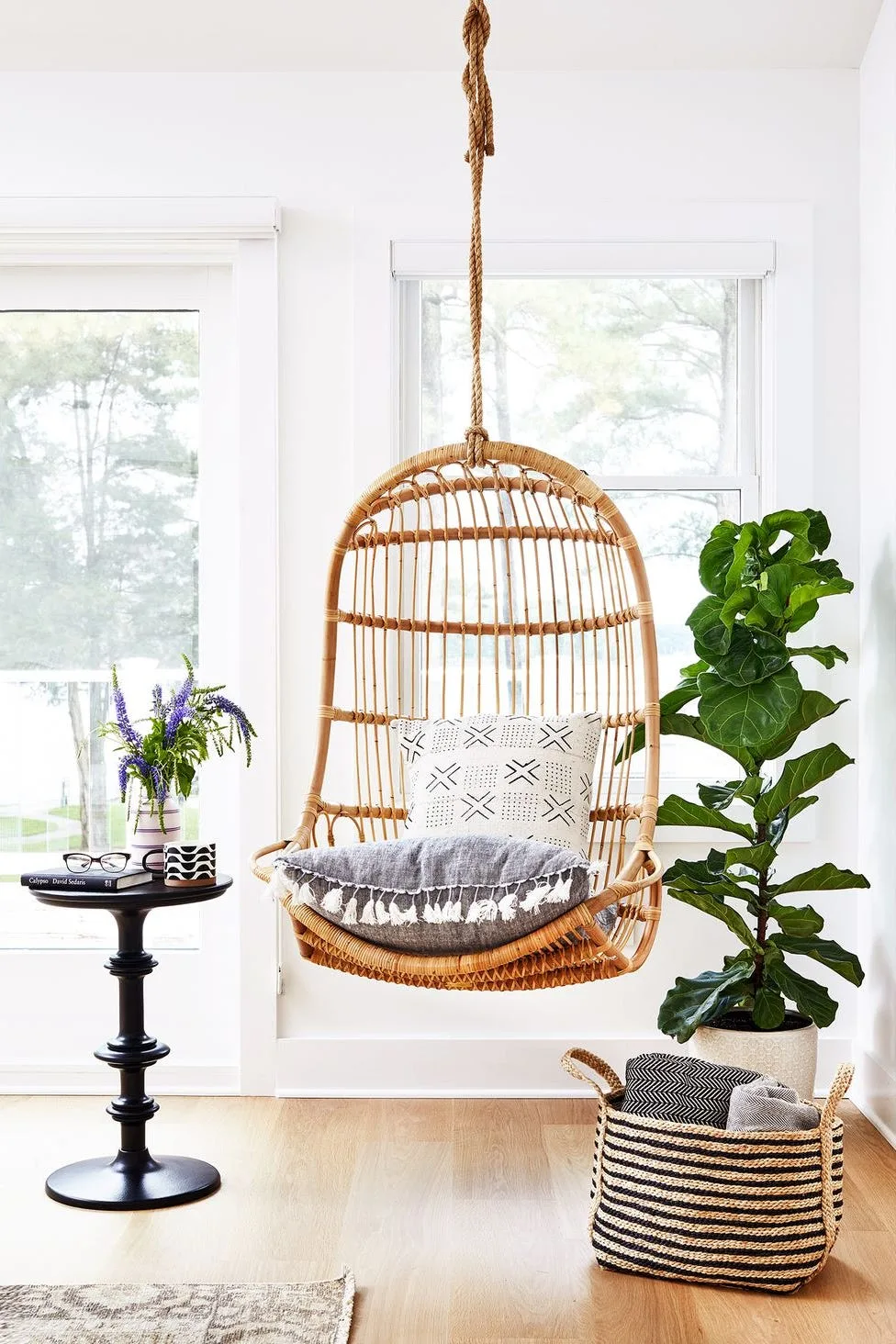 Bright bedroom sitting corner with woven hanging chair, tall broad-leaved plant, small dark round table, two windows and basket of folded blankets.