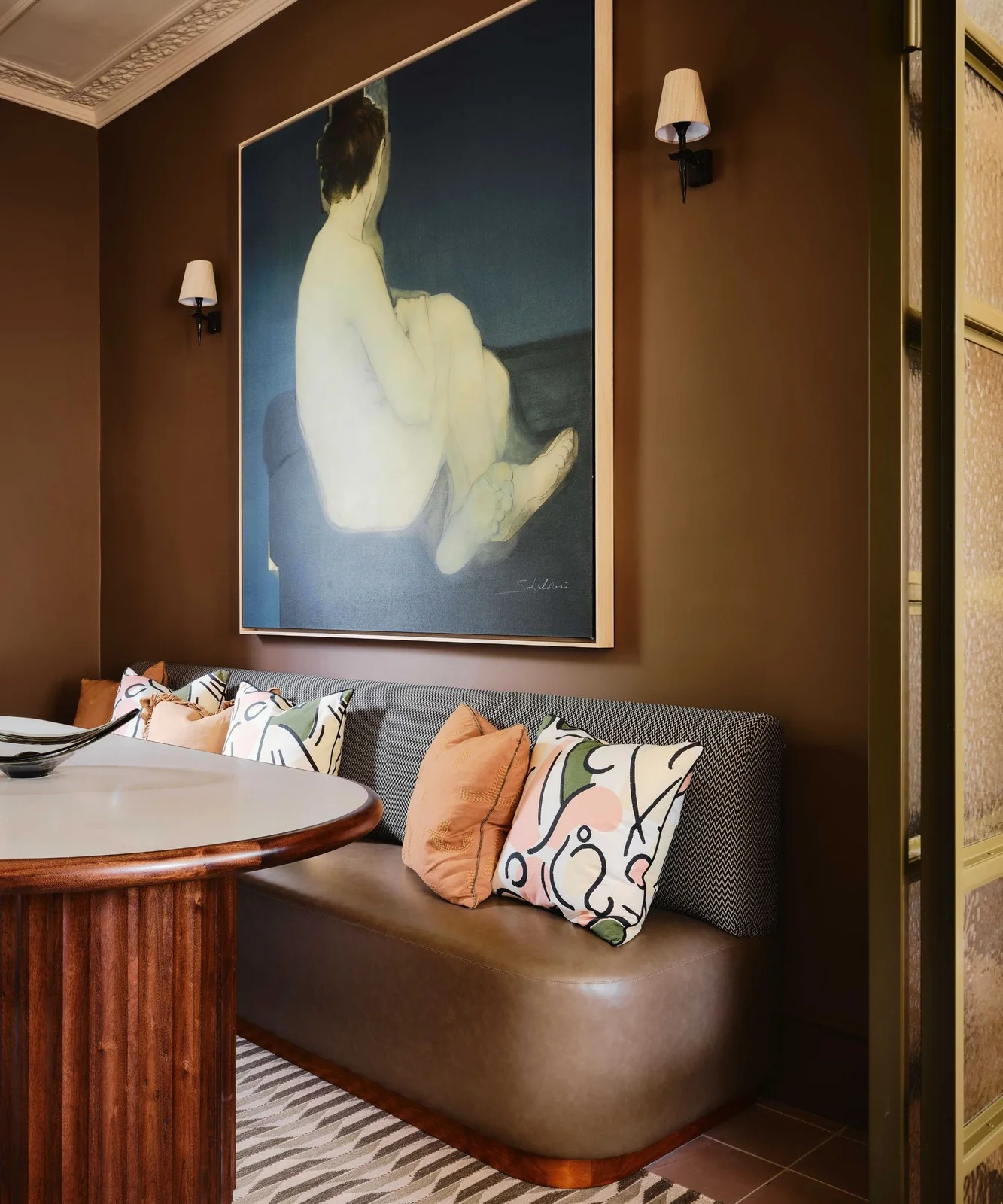 A curved dining bench with a finely patterned back carries graphic and peach cushions beneath a large portrait beside a fluted table pedestal
