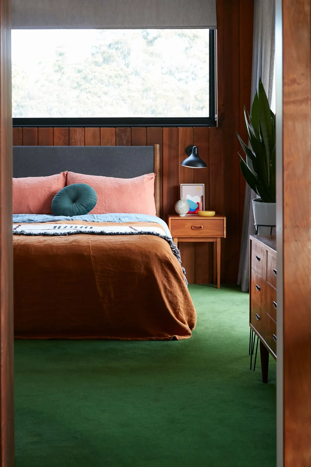 Green bedroom carpet runs beneath a bed with orange and pink bedding beside wood wall panels, a small wood nightstand and high window