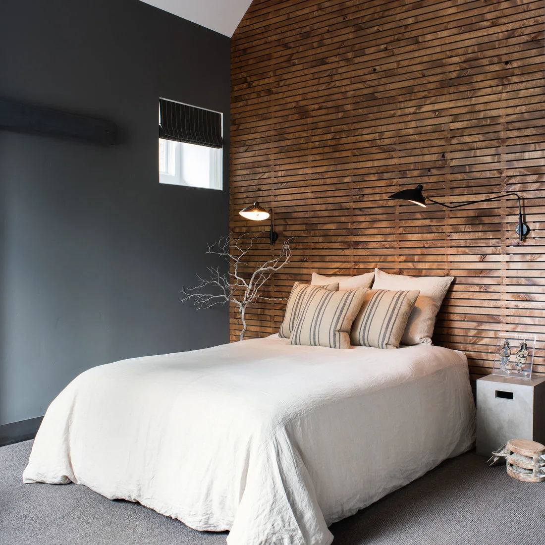 Pale bedding and striped cushions sit before a wooden slat wall with two black adjustable lamps beside a charcoal wall and small window