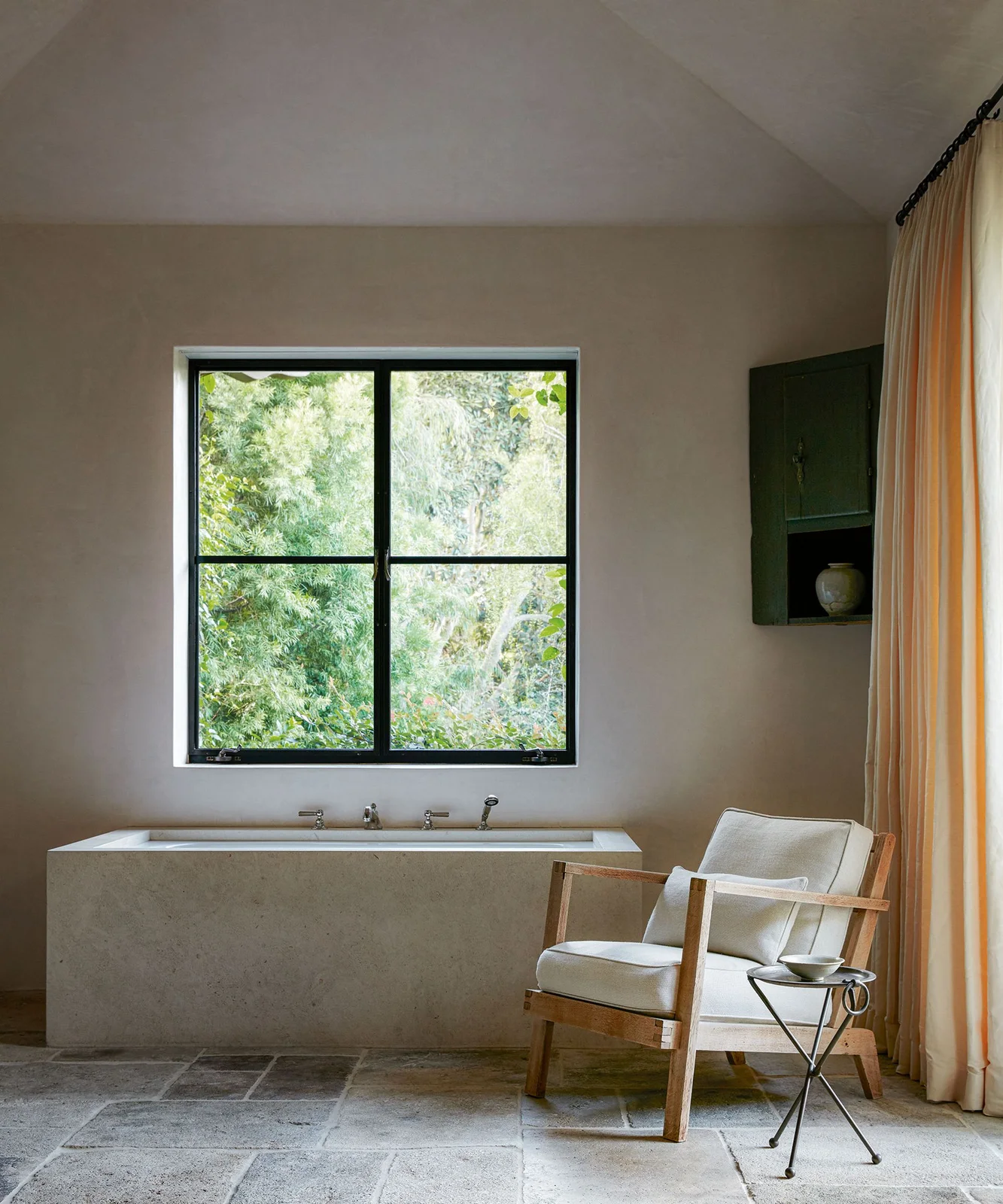 A pale rectangular bath sits beneath a dark-framed window beside a wooden armchair, cream curtains and a small round table