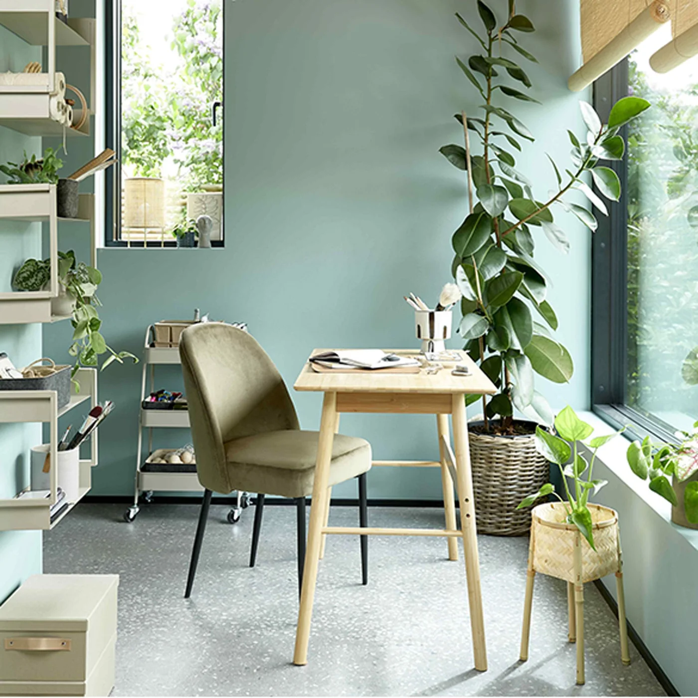 Pale green walls surround a small wooden desk, upholstered chair, open shelving and several leafy plants