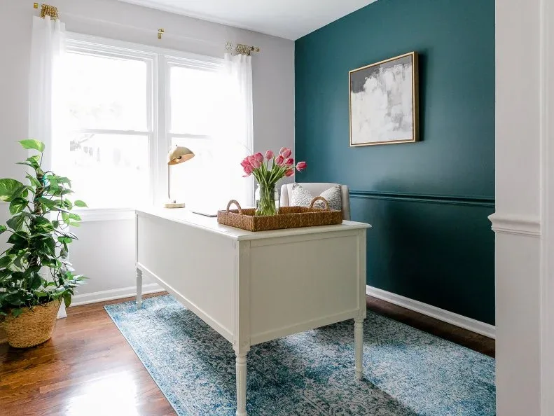 A pale freestanding desk sits beside a teal-green paneled accent wall and light curtains beneath a warm metal picture frame