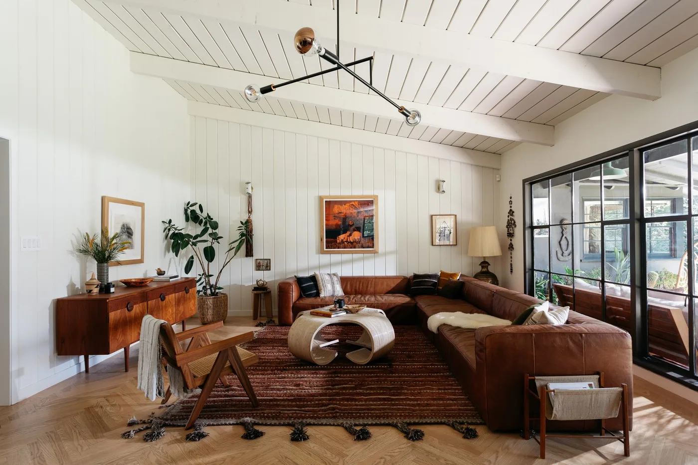 Brown sectional living room with white board walls, wood sideboard, warm striped rug and black-framed glazing