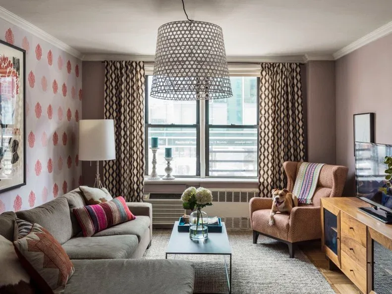 Living room with gray sectional, warm patterned wing chair, pink wall motifs, dark patterned curtains, narrow table and wooden media cabinet.