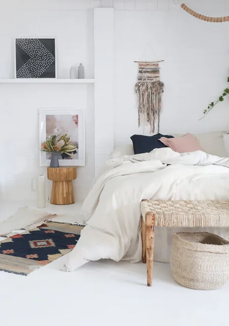 A woven wall hanging appears above a pale bed with a timber bench nearby.