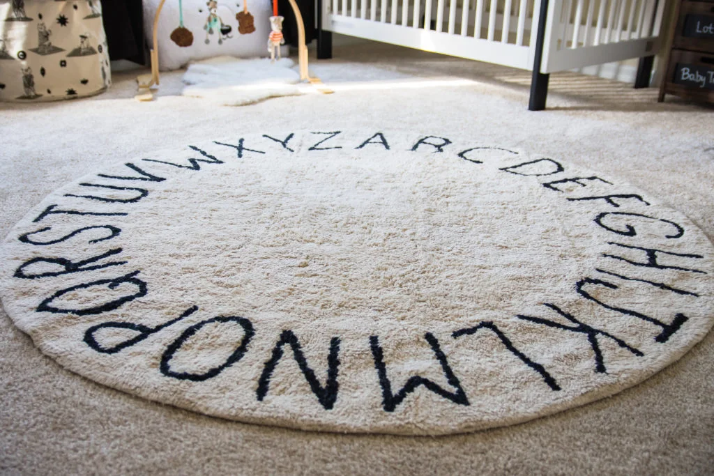 A pale circular nursery rug with dark letters sits on light carpet near patterned storage containers and a separate play arch