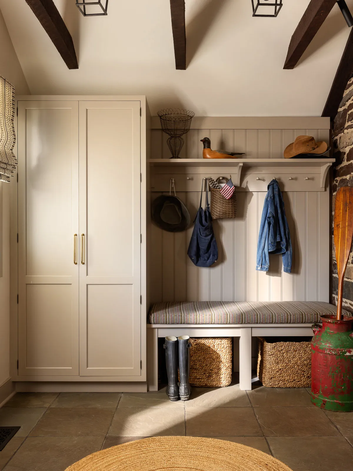 A pale bench and coat hooks sit beneath exposed timber beams beside a tall cupboard.