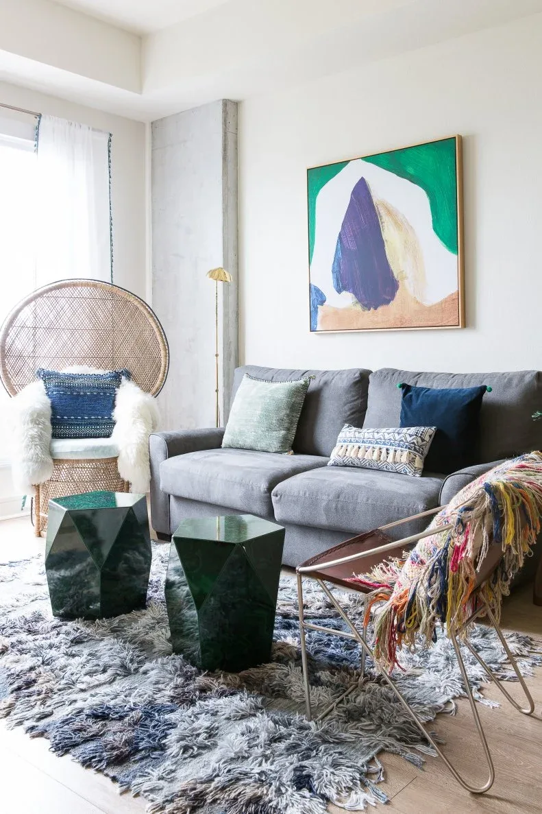 Living room with gray sofa, thick textured gray rug, woven high-backed chair, dark green faceted tables and abstract green-and-purple artwork.