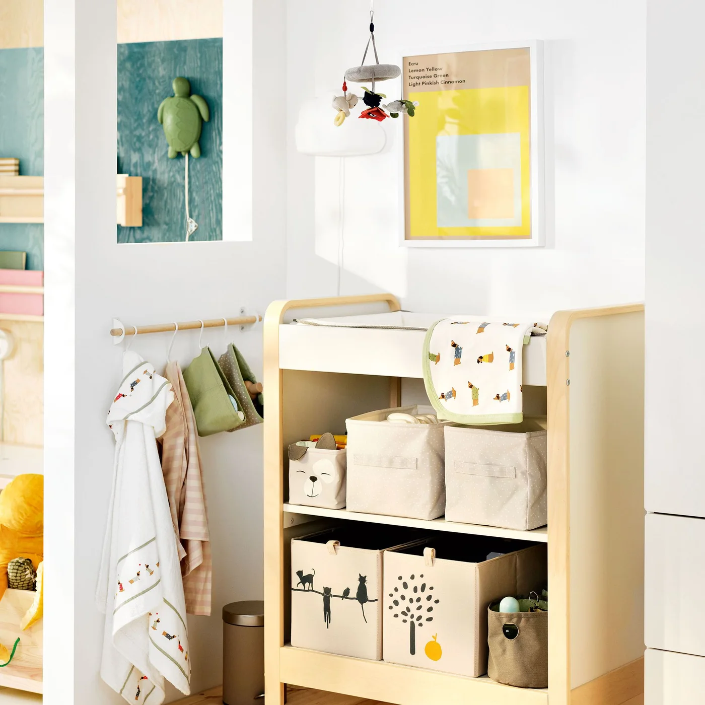 A pale wood changing unit holds fabric bins beneath a light changing surface beside white walls, a small clothes rail and a framed yellow picture
