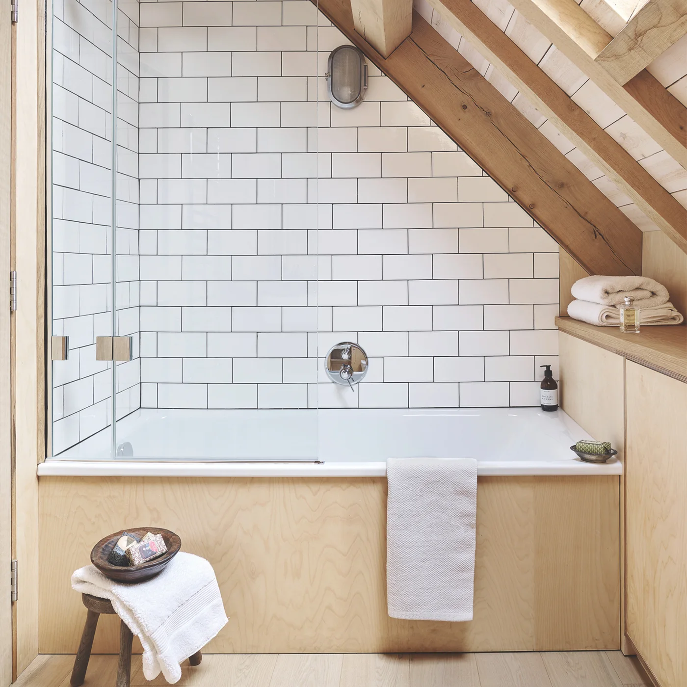 A white bath beneath exposed wooden roof framing is surrounded by white rectangular tiles beside a glass screen, pale wooden panels and a small stool