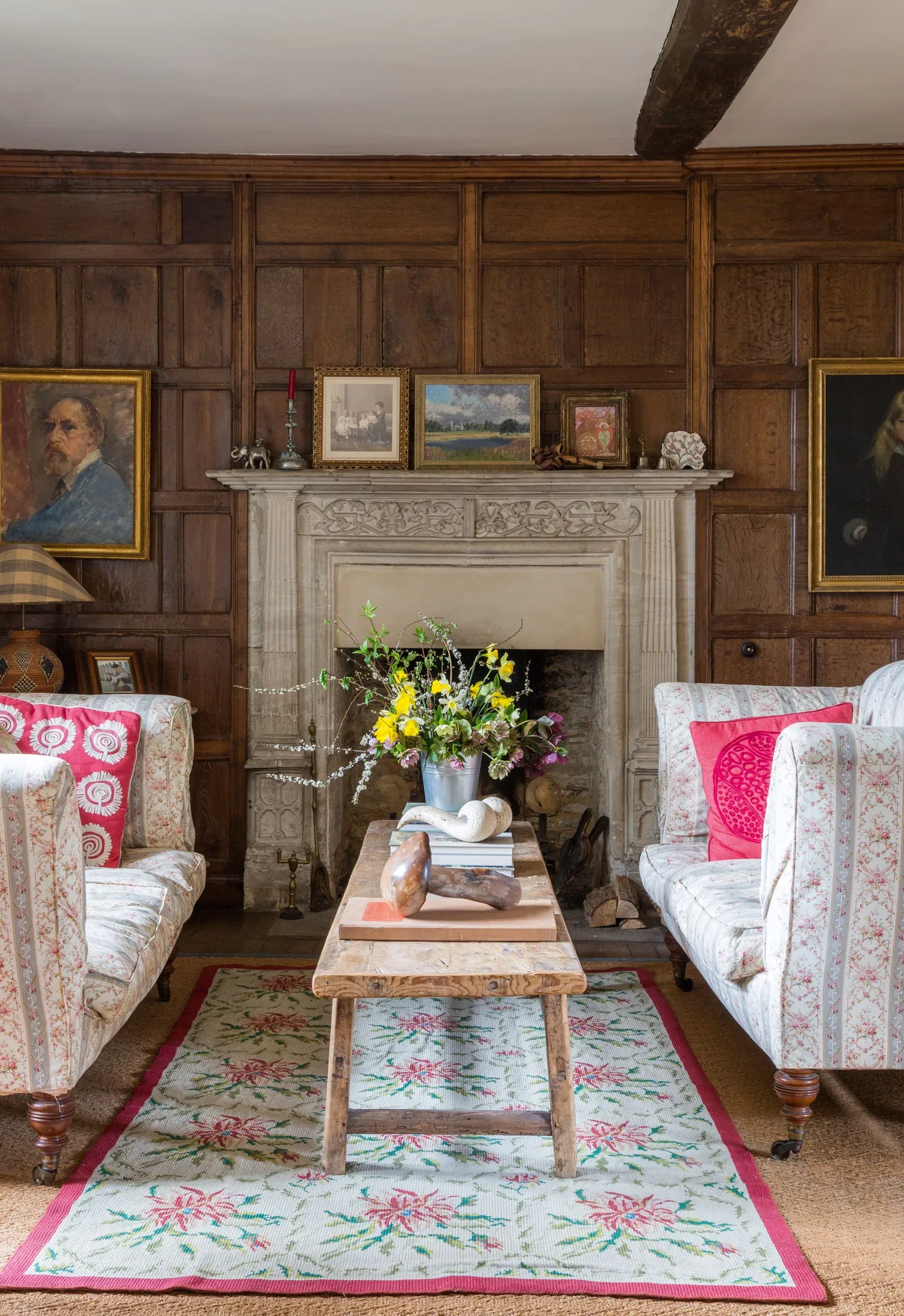 Pale carved fireplace against dark wooden panels with floral armchairs, flower-patterned rug and framed pictures.