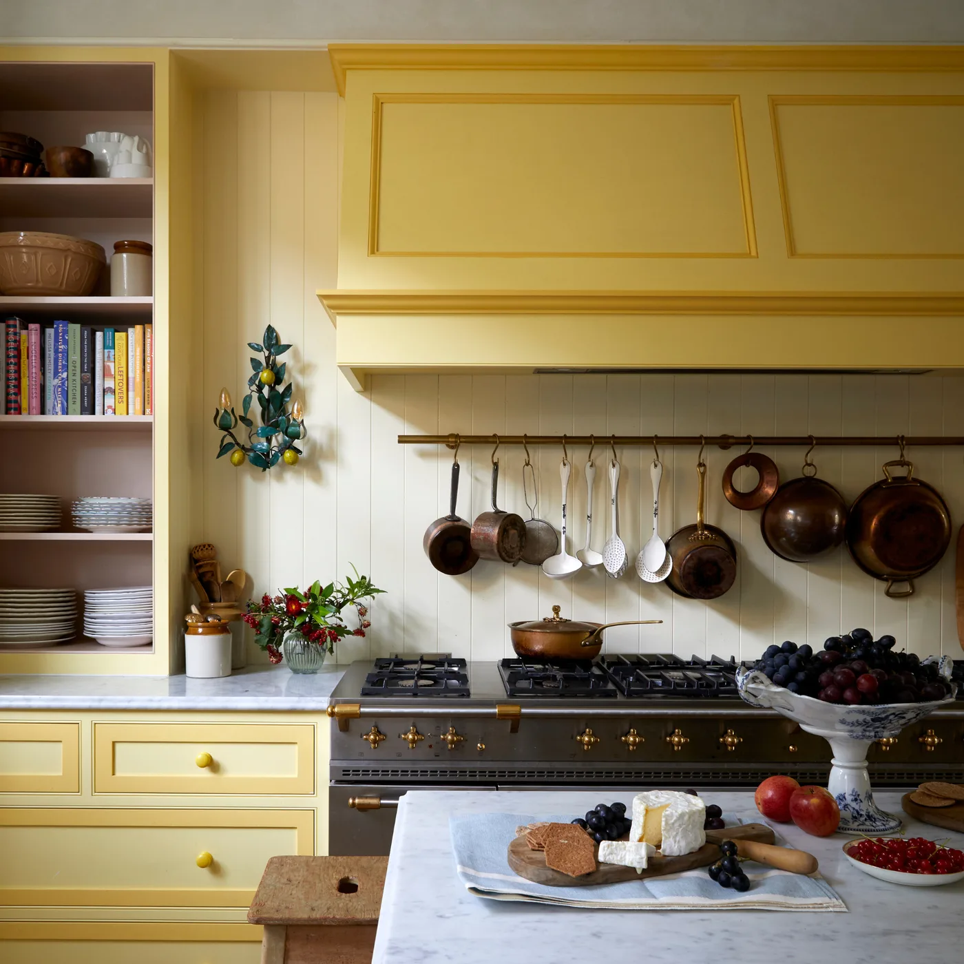 Yellow paneled kitchen with a matching hood, dark range cooker, hanging pans and an open shelf of dishes and books.