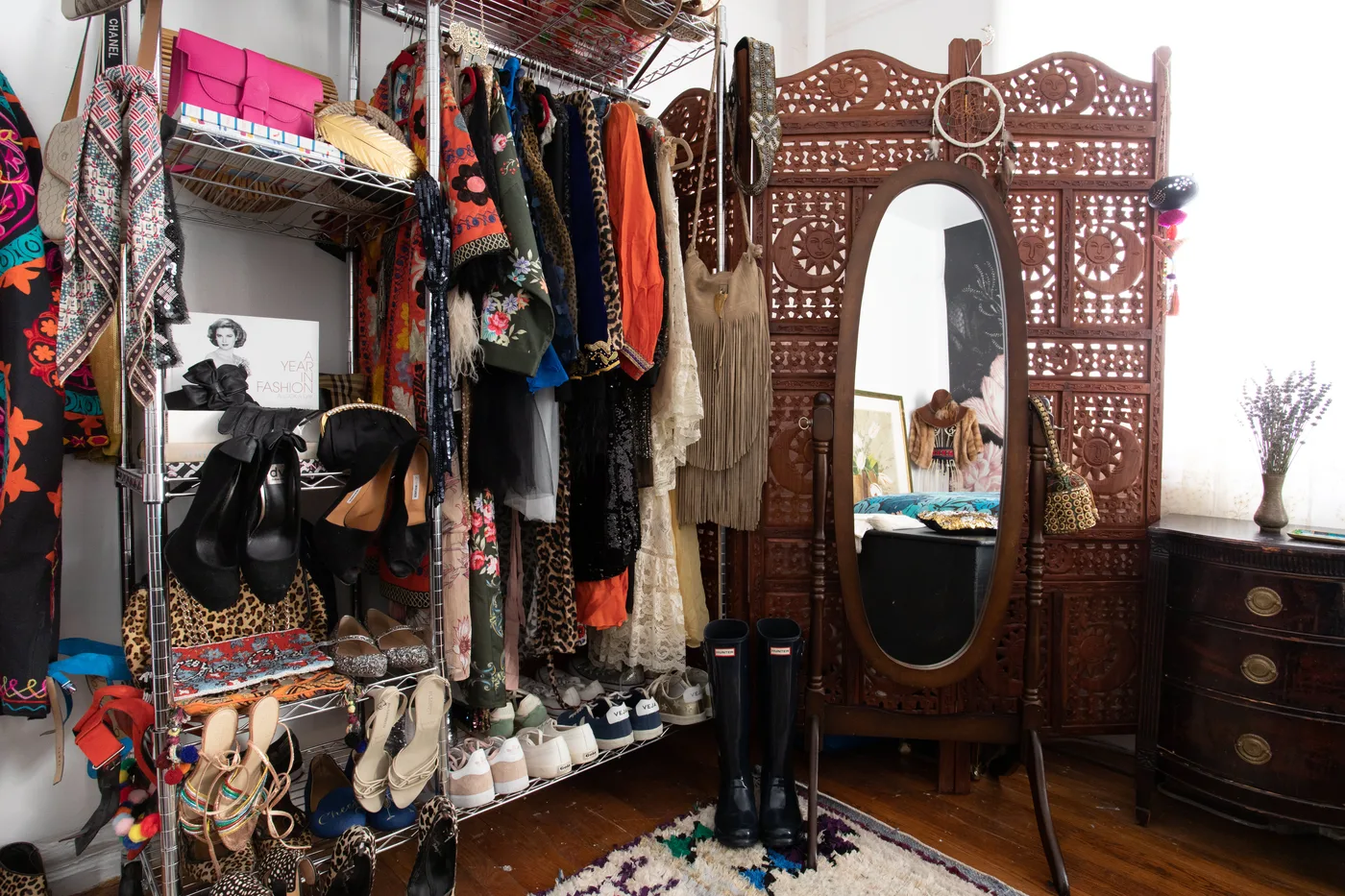 Bedroom dressing area with carved wooden screen and oval mirror, open clothes racks, baskets, patterned garments, dark drawers and colorful rug.