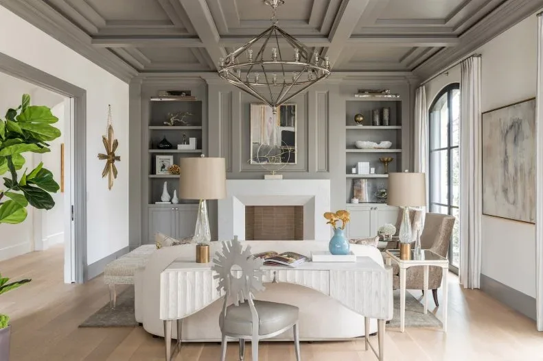 Living room with gray coffered ceiling, white ribbed sofa, pale wooden-framed chairs, gray ottoman, white fireplace and gray built-in shelves.