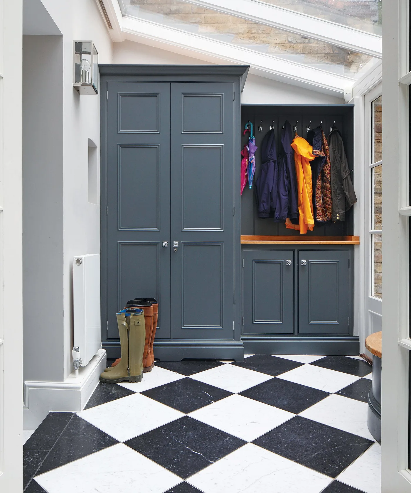 Blue-gray mudroom with tall closed cabinet beside open coat hooks and wooden seat under a sloping glazed roof.