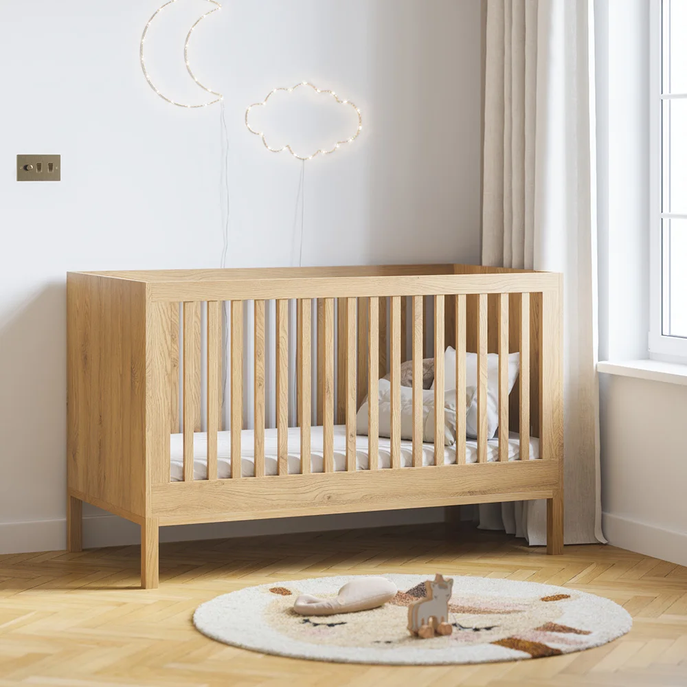 A plain wooden crib stands against a pale wall beside a window, small round rug and decorative wall lights