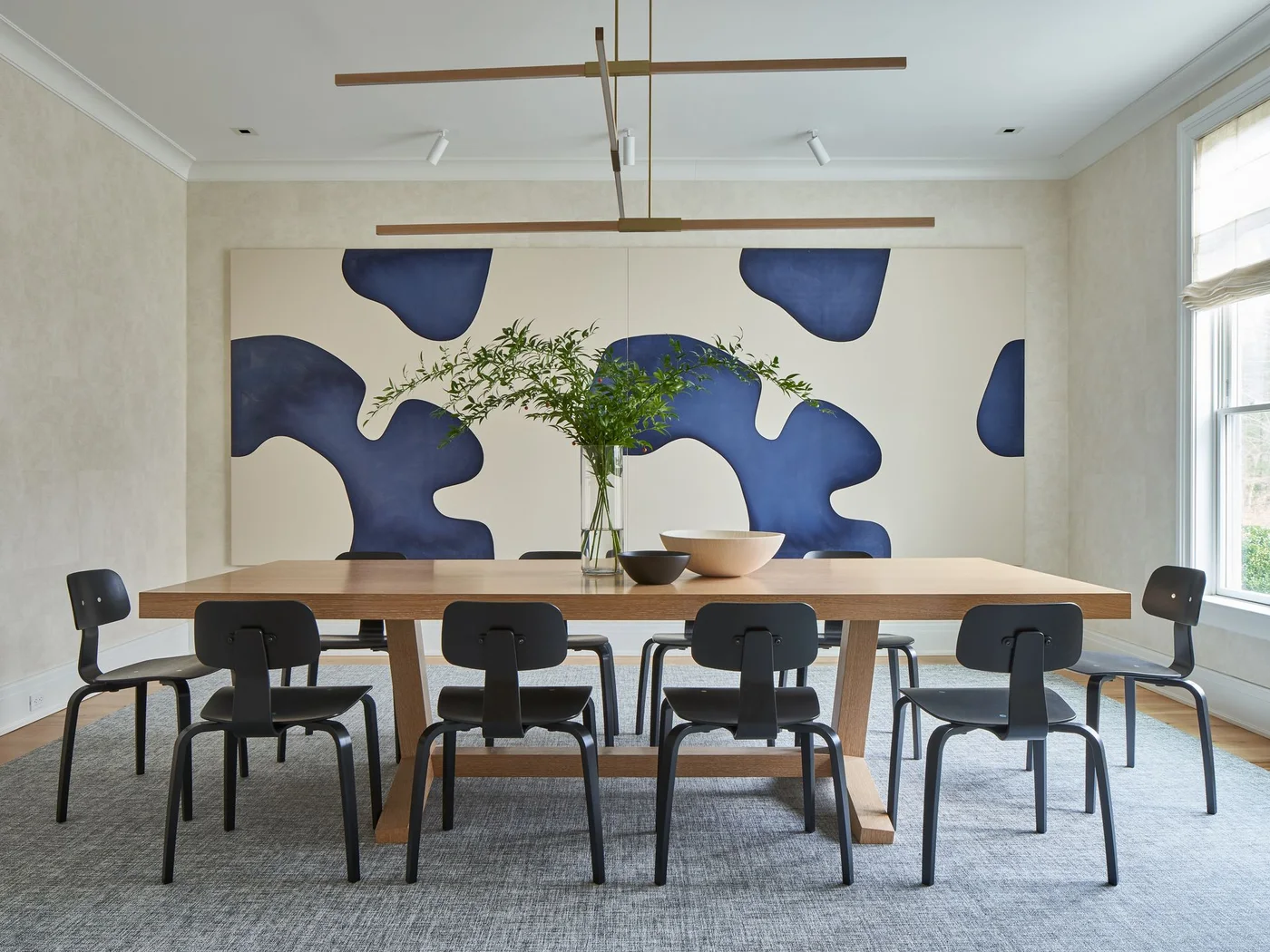 A rectangular wood dining table and black chairs stand beneath a narrow linear light in front of a large blue abstract artwork against pale walls