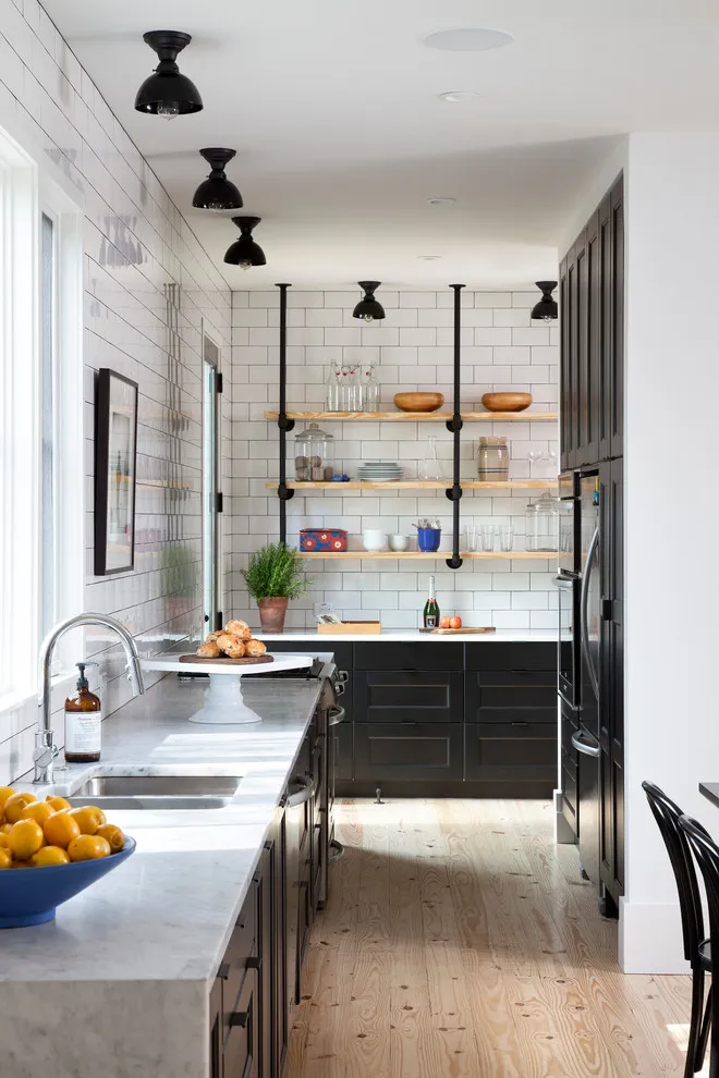 Dark lower cabinets and tall storage sit beneath white tiled walls and open timber shelves in a narrow kitchen