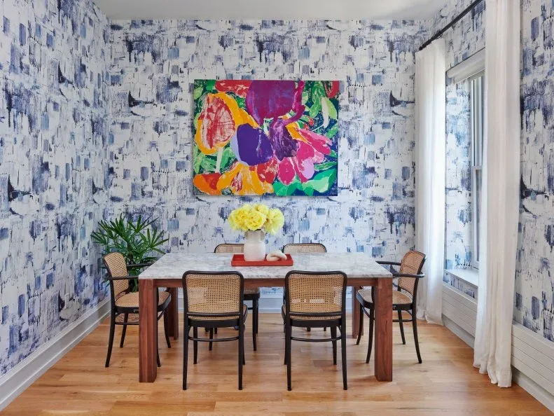 Plain white curtain beside a window in a dining room wrapped in blue-and-white abstract wallpaper, with colorful artwork, a wooden table, and woven-back chairs.