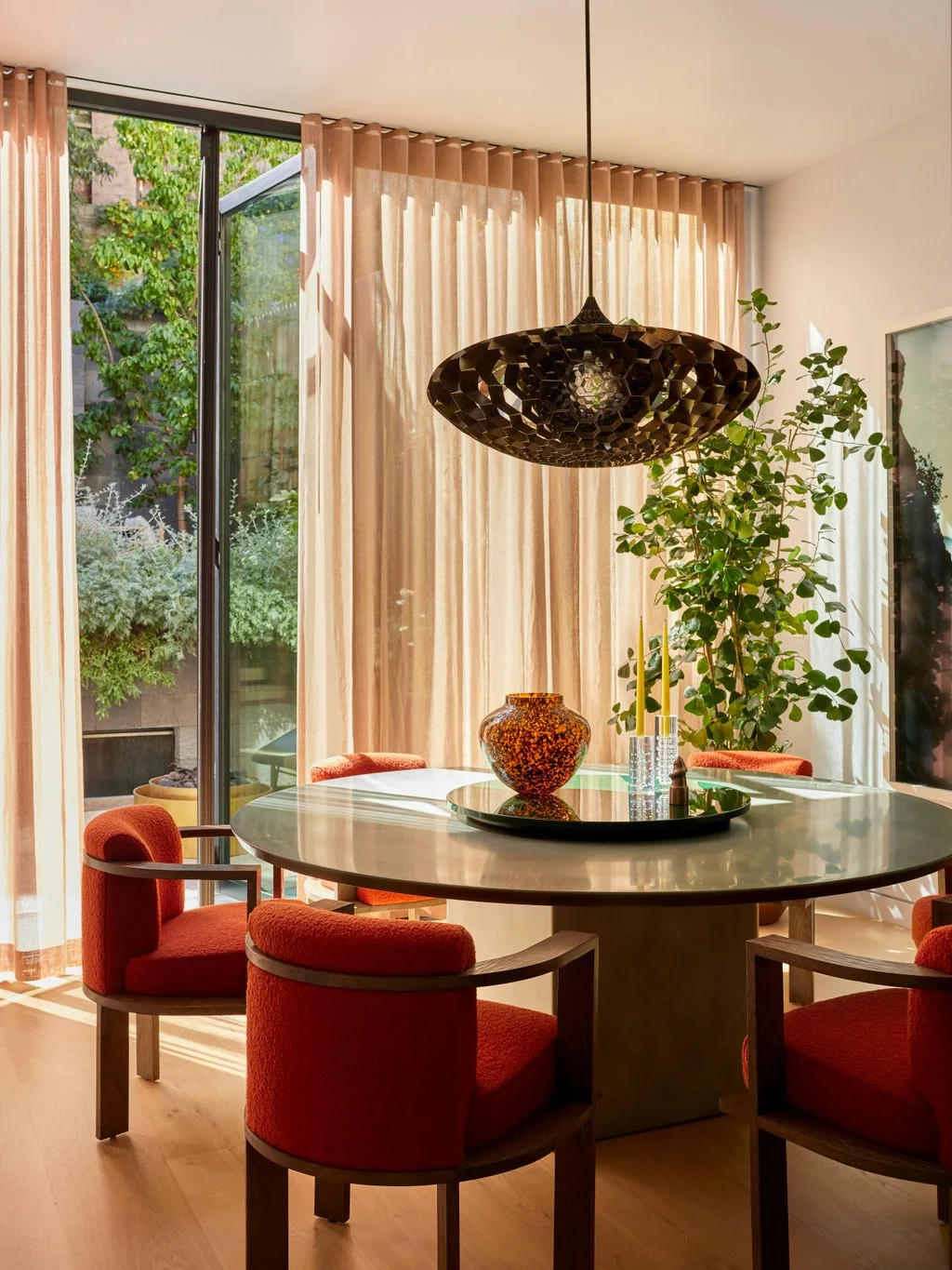 Orange upholstered dining chairs surround a reflective round table beneath a dark sculptural pendant beside sheer pale curtains