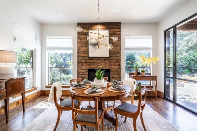 Dining room with a wood-paneled fireplace wall, round wood table, curved chairs, pale window shades and branching globe light