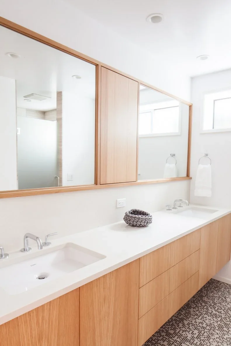 A long pale wood double vanity stands below two mirrors divided by a central wood cupboard beside white walls and a small patterned bathroom floor