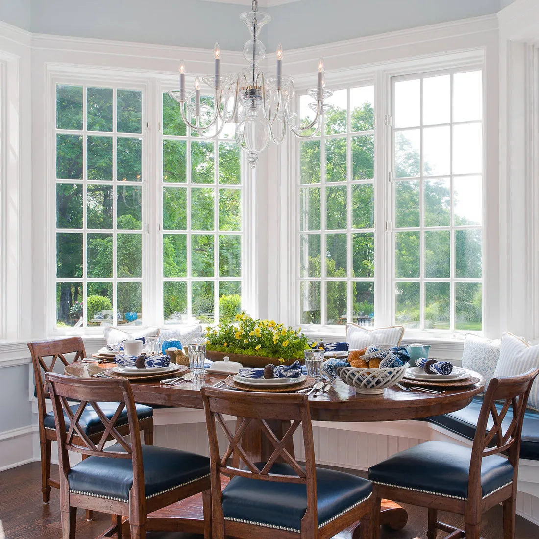 Wood oval dining table with cross-back chairs, blue cushions and place settings in a glazed bay