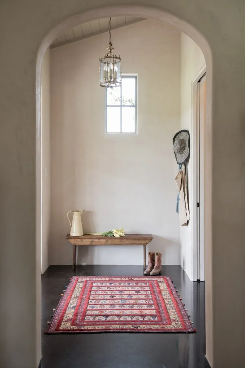 Clear candle lantern inside arched entry with high window, simple wood bench, red patterned rug and dark floor
