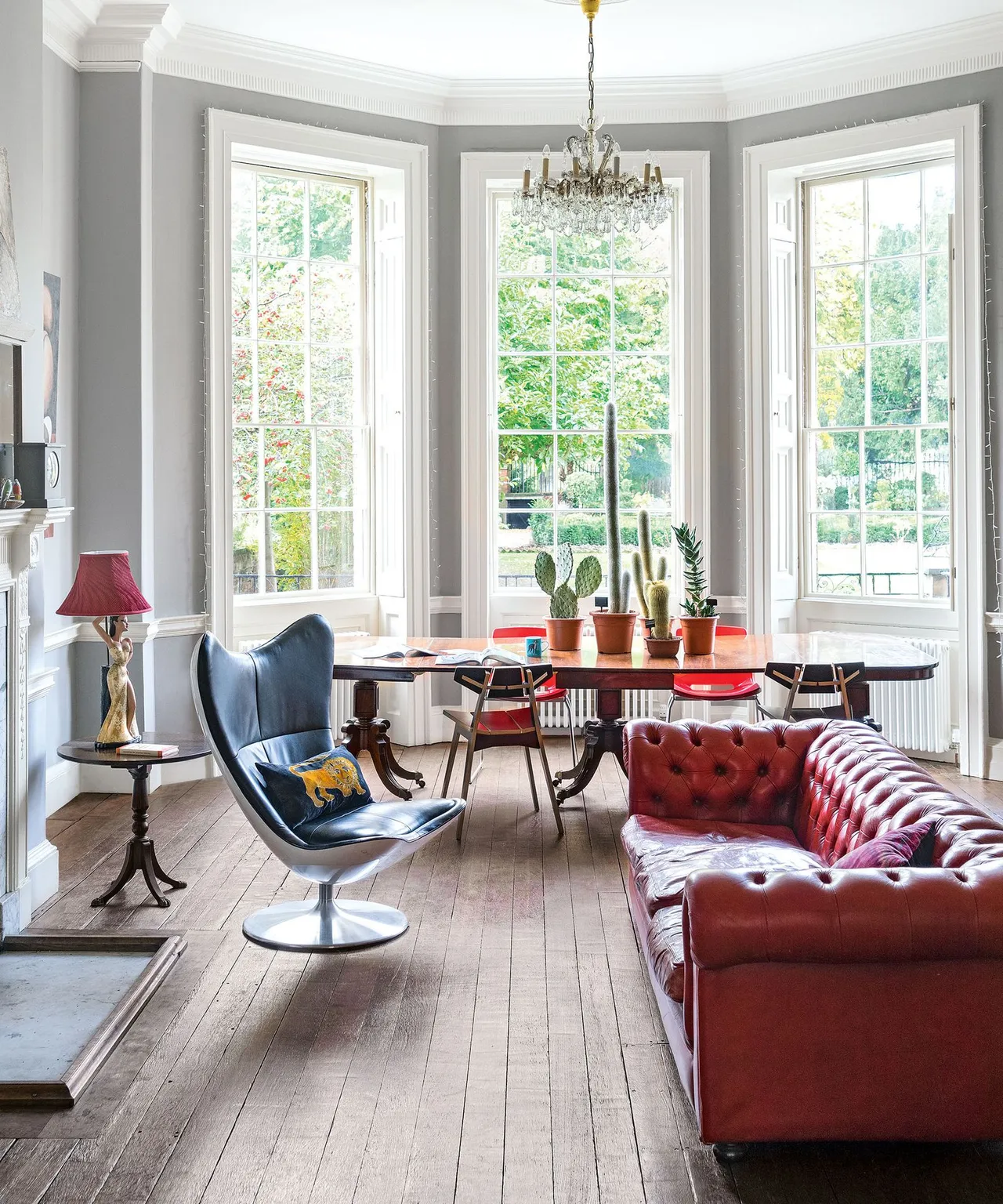 Living room with red buttoned sofa, blue wing-shaped chair, pale gray walls, tall windows, wooden floor and a table with potted plants beyond the seats.