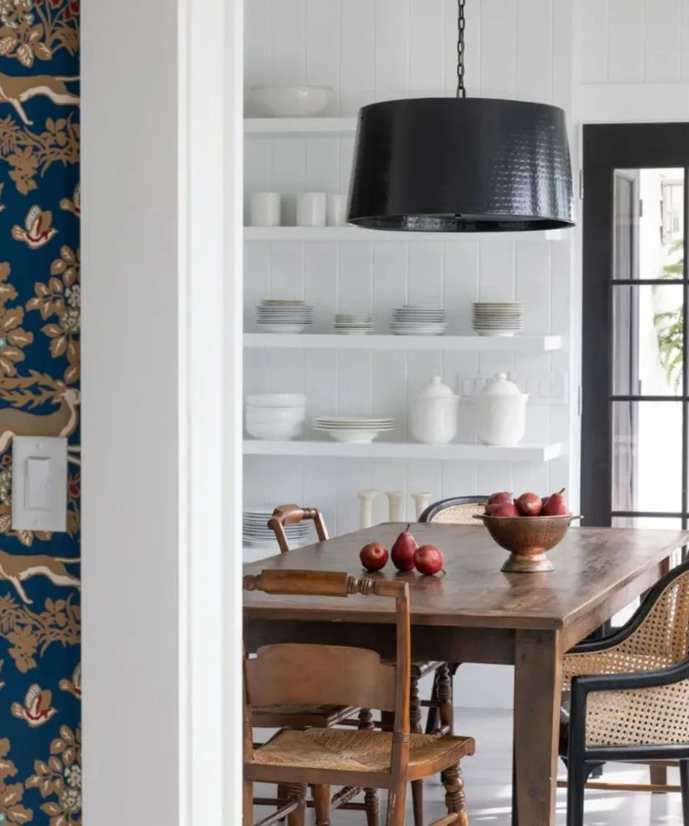 Cottage dining corner with a wooden table, mixed wooden and dark-framed chairs, white open shelves displaying pale ceramics and a dark pendant shade.