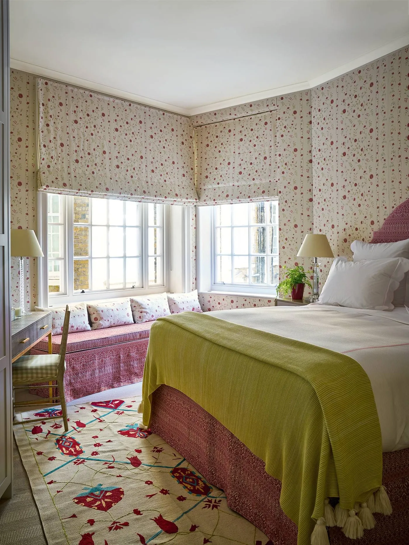 Fine pink floral wallpaper and matching blinds around a bedroom window seat with pink cushions.