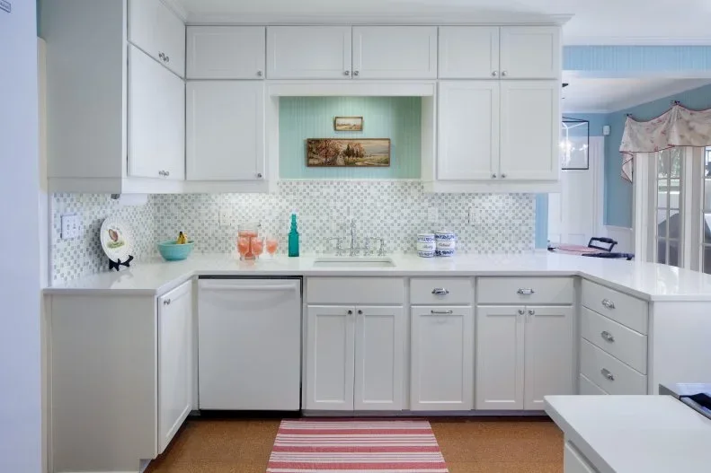 Pale kitchen cupboards with simple knobs stand above a small patterned backsplash, a sink and darker-front appliances beside a striped floor runner