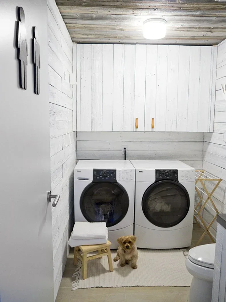 Compact white laundry room with board walls, weathered-looking ceiling, side-by-side machines, upper cabinets and small drying rack