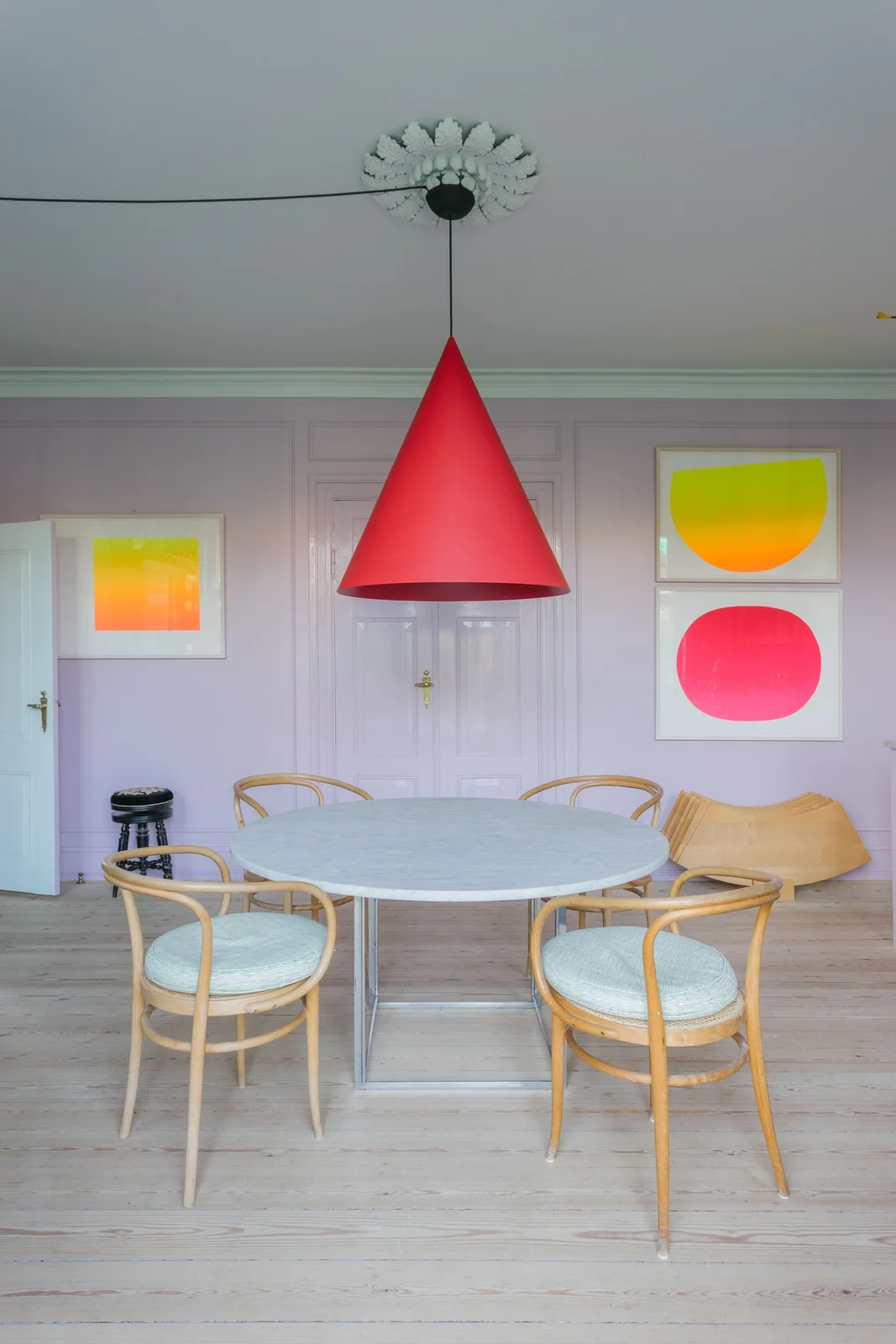 A pale round dining table and curved light wood chairs sit beneath a red cone pendant beside lilac paneled walls and bright geometric pictures