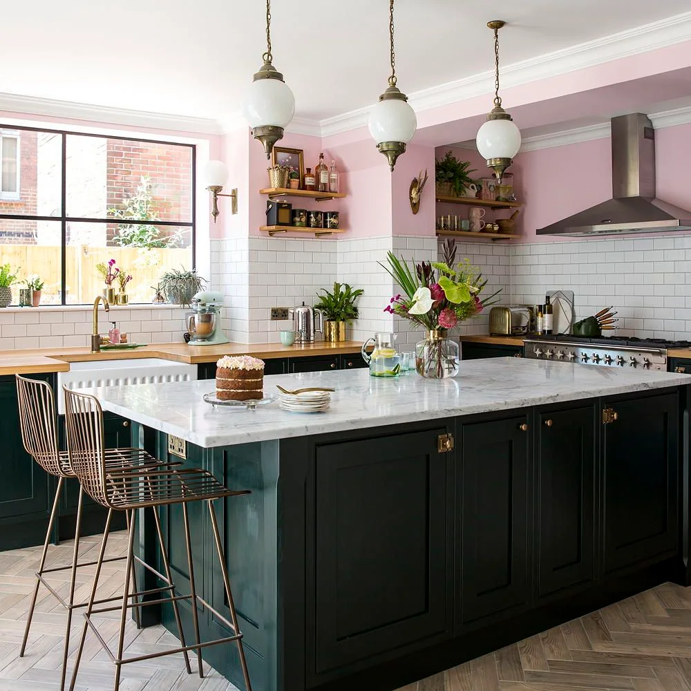 White offset tiles line a kitchen beneath pink walls and wooden shelves beside a dark green island and pale counter