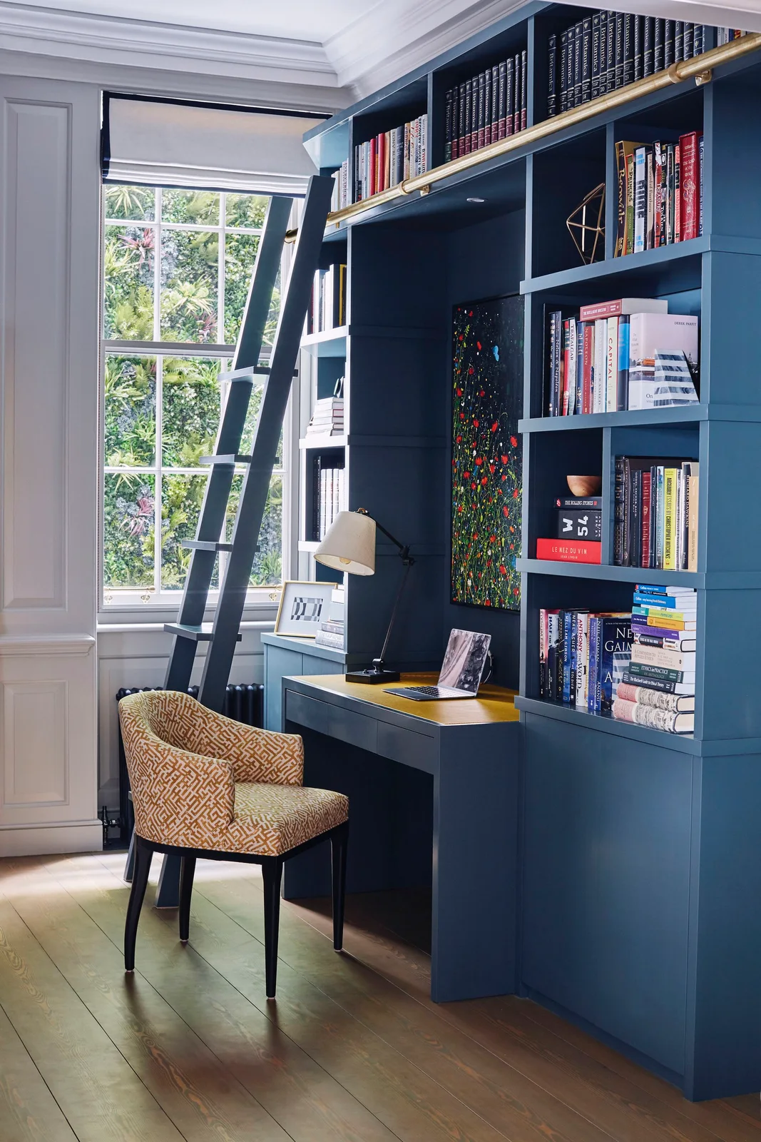 Blue bookcases and a fitted desk sit beside a tall window with a patterned chair, adjustable lamp and dark library ladder